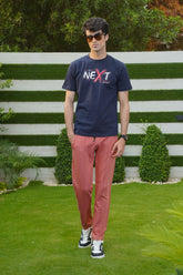 A person wearing a navy blue graphic t-shirt with the word 'NEXT' printed on the front, paired with red pants and standing in front of a white fence.